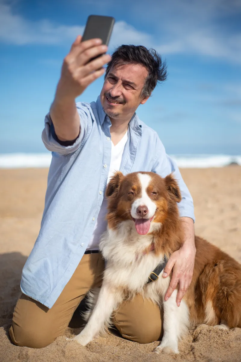 Man maakt op het strand een selfie met een bruin-witte hond naast zich.